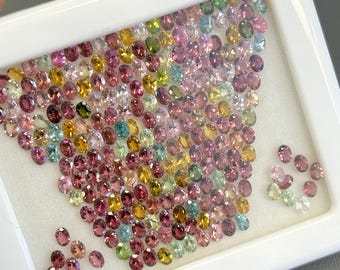May include: A collection of colorful oval-shaped gemstones in a white display tray. The gemstones are a mix of pink, green, yellow, and clear.