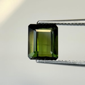May include: A green gemstone with a rectangular emerald cut. The gemstone is held in a pair of tweezers.