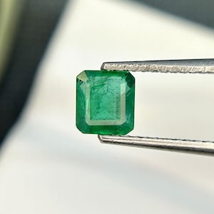 Puede incluir: Una gema de esmeralda verde vibrante, tallada en forma de esmeralda, se sujeta con unas pinzas sobre un fondo blanco.