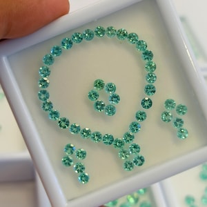 Può includere: Una collezione di piccole gemme rotonde color verde acqua disposte in un motivo decorativo all'interno di un vassoio bianco. Le gemme sono disposte a spirale e a motivo floreale, con pietre sciolte aggiuntive.