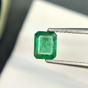 Puede incluir: Una esmeralda verde vibrante, tallada en forma cuadrada, se sostiene con unas pinzas. La esmeralda tiene un aspecto ligeramente nublado, con inclusiones visibles.