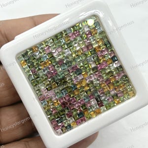 Könnte beinhalten: Ein weißer quadratischer Behälter gefüllt mit zahlreichen kleinen, quadratisch geschliffenen Edelsteinen. Die Edelsteine weisen eine Vielzahl von Farben auf, darunter Rosa, Grün, Gelb und Blau. Der Behälter wird in einer Hand gehalten, und die Edelsteine sind dicht gepackt.