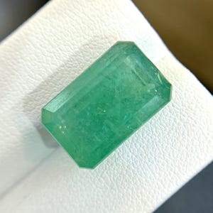 Puede incluir: Una gema de esmeralda verde vibrante tallada en forma de esmeralda. La gema está facetada y tiene una apariencia translúcida.