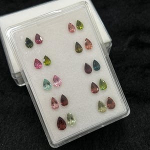 Könnte beinhalten: Eine transparente Plastikbox gefüllt mit 12 birnenförmigen Edelsteinen. Die Edelsteine sind paarweise angeordnet und haben verschiedene Farben, darunter Grün, Rosa, Blau und Braun.