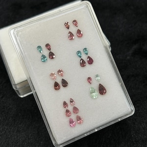 May include: A white box with a clear lid containing 12 pear-shaped gemstones. The gemstones are arranged in rows and are various shades of pink, green, and blue.