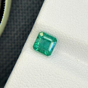 Może przedstawiać: Żywy, szmaragdowy zielony kamień szlachetny. Kamień jest osadzony na białej, teksturowanej powierzchni. Fasetki kamienia szlachetnego odbijają światło, ukazując jego przejrzystość i kolor. Obraz zawiera również fragment zegarka.