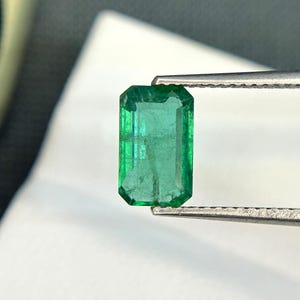 Puede incluir: Una gema de esmeralda verde vibrante, tallada en forma de esmeralda, se sostiene con unas pinzas. La gema es transparente y tiene una ligera forma rectangular.