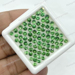 May include: A white tray filled with 100 round, green gemstones. The tray is held in a hand.