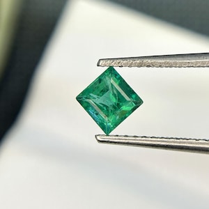 Puede incluir: Una gema de esmeralda verde vibrante, cortada en forma cuadrada, se sostiene con unas pinzas. La esmeralda es transparente y tiene un ligero brillo.