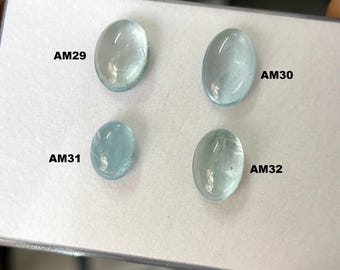 Op de afbeelding: Vier ovale, lichtblauwe edelstenen worden gepresenteerd op een wit oppervlak. Elke edelsteen is gemarkeerd met zwarte tekst: AM29, AM30, AM31 en AM32. De edelstenen hebben een doorschijnend uiterlijk, met subtiele interne patronen. De presentatie is netjes en professioneel.