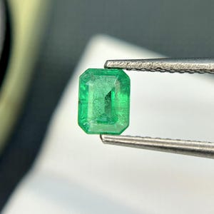 Puede incluir: Una esmeralda verde vibrante, tallada en forma rectangular, se sostiene con unas pinzas.
