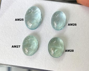 Op de afbeelding: Vier ovale aquamarijn edelstenen in verschillende blauwgroene tinten. Elke steen is gelabeld met een code: AM25, AM26, AM27 en AM28. De edelstenen worden gepresenteerd op een witte ondergrond.