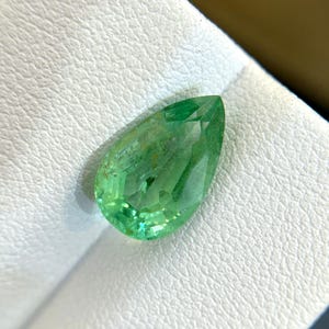 Puede incluir: Una piedra preciosa verde en forma de pera con facetas. La piedra preciosa está sobre una superficie blanca.