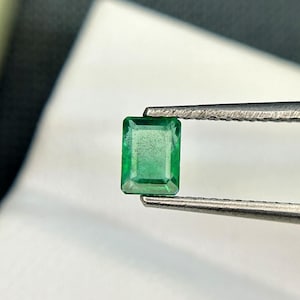 Puede incluir: Una esmeralda verde vibrante, tallada en forma rectangular, se sostiene con unas pinzas.