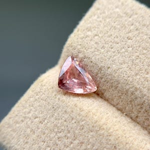 May include: A faceted, triangular gemstone with a soft pink hue. The gem is set against a beige, textured background, showcasing its polished surfaces and internal clarity. The gem's shape is a perfect triangle.
