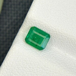 Puede incluir: Una gema verde de talla esmeralda y forma rectangular. La gema tiene bordes biselados y un color verde intenso. La piedra se encuentra sobre una superficie blanca texturizada.