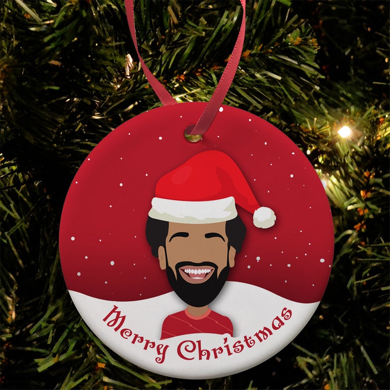 Pu&ograve; includere: Un addobbo natalizio rotondo rosso e bianco con un'illustrazione di un uomo con un cappello di Babbo Natale e una maglia rossa e bianca. L'addobbo dice "Buon Natale" in un font bianco corsivo.