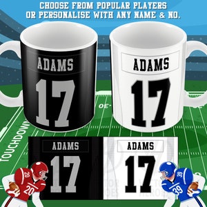 May include: Two white ceramic mugs with black handles. The mugs are personalised with the name "Adams" and the number "17". One mug has a black background and the other has a white background. The mugs are shown on a green and white football field.