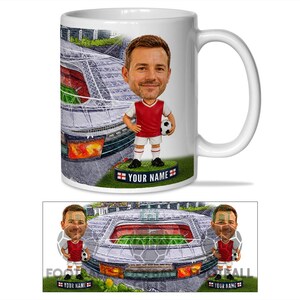 May include: A white ceramic mug featuring a graphic of a football stadium and a cartoon-style figure wearing a red and white football kit, holding a football. The figure stands on a base with the text YOUR NAME flanked by two small English flags.