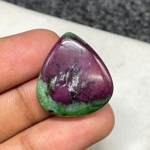 Könnte beinhalten: Ein polierter Edelstein in Tropfenform. Der Stein hat eine tief burgunderrote Mitte mit weißen und grauen Einschlüssen, die in einen leuchtend grünen Rand übergehen. Der Edelstein wird in einer Hand gehalten.