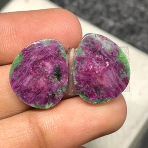 May include: Two oval-shaped ruby zoisite stones. The stones feature a vibrant magenta center with green edges. The stones are held together with clear tape. The stones are displayed against a neutral background.