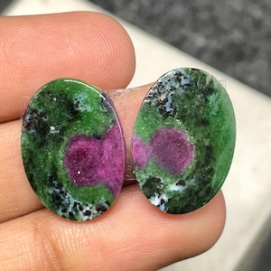 May include: Two oval-shaped gemstones. Each stone features a vibrant mix of colors: a central, deep magenta hue surrounded by a rich green, with accents of black and white. The stones are held in a hand.