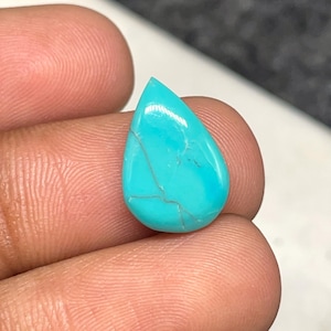May include: A teardrop-shaped turquoise gemstone with a polished surface. The stone is a vibrant blue-green color with natural veining. The gemstone is held between fingers.