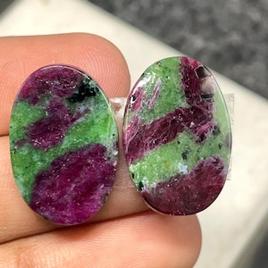 May include: Two oval-shaped gemstones with a vibrant mix of colors. The stones feature a striking combination of deep red and green hues, with some black inclusions. The polished surfaces showcase a natural, organic pattern, perfect for jewelry.