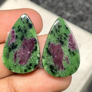 May include: Two teardrop-shaped gemstones. The stones have a green base with black speckles and large, irregular patches of deep red. The gems are polished and have a glossy surface, suitable for jewellery making.