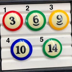 Może przedstawiać: Kolekcja pięciu okrągłych guzików w stylu kul bilardowych. Każdy guzik ma kremowy środek z czarnym numerem i kolorową obwódką: czerwoną (3), zieloną (6), żółtą (9), niebieską (10) i zieloną (14).