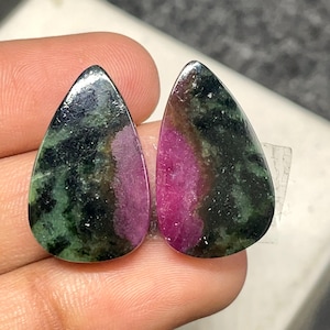 Może przedstawiać: Dwa wypolerowane kamienie szlachetne w kształcie łezki. Kamienie charakteryzują się mieszanką kolorów, w tym głęboką zielenią, czernią i żywym różem. Kamienie szlachetne są gładkie i mają błyszczące wykończenie, co sugeruje, że są gotowe do tworzenia biżuterii lub ekspozycji.