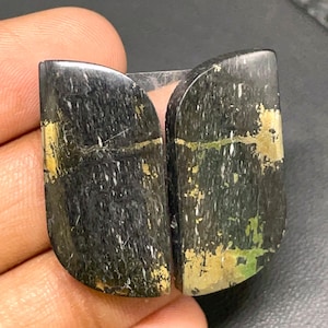 May include: Two black and green cabochons with gold flecks. The cabochons are shaped like a teardrop and have a smooth, polished surface.