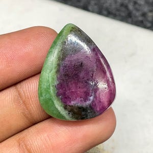 Könnte beinhalten: Ein polierter, tropfenförmiger Edelstein mit einer lebendigen Farbpalette. Der Stein hat eine satte, tiefmagentafarbene Mitte, die in ein dunkelgraues Band übergeht und von einem hellgrünen Rand umgeben ist. Der Edelstein wird in einer Hand gehalten.