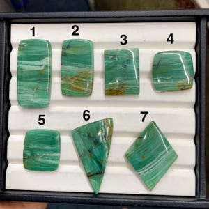 以下が含まれることがあります： 長方形、正方形、三角形など、さまざまな形状の7つの研磨された宝石のコレクション。石は鮮やかな緑色で、白と茶色の自然な模様があり、黒いトレイに入っています。