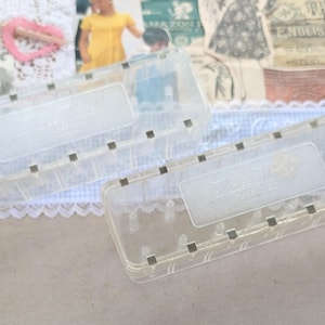 May include: Two rectangular, clear plastic storage containers with hinged lids. Each container features a series of small, evenly spaced metallic squares along the front edge and a rectangular embossed label area on the lid surface.