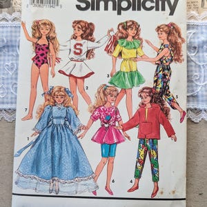 May include: A Simplicity 7600 sewing pattern envelope featuring illustrations of seven dolls wearing various outfits, including a blue gown, a cheerleader uniform, a pink star-print top, and a red jacket with patterned pants.