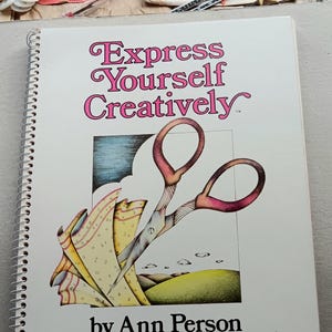 May include: A spiral-bound book titled "Express Yourself Creatively" with a colorful illustration of scissors cutting fabric. The cover features the author's name, Ann Person, and is surrounded by sewing notions.