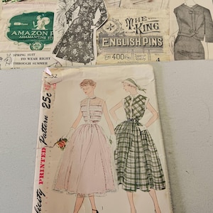 May include: Vintage sewing patterns and ephemera are arranged on a light-colored surface. The patterns feature illustrations of dresses, including a pink dress and a green plaid dress. Other items include pins, buttons, and a ruler.