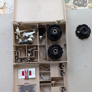 May include: Tan sewing machine accessory kit with various parts, including bobbins, presser feet, and a brush. The kit includes a Singer 2020 needle pack. The kit is open, revealing the contents.