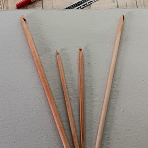 May include: Four wooden crochet hooks of varying lengths and shades of brown are arranged on a light gray surface. The hooks are slender, with a hook at one end. Other craft supplies, including buttons and sewing patterns, are visible in the background.