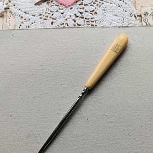 May include: A vintage crochet hook with a cream-colored handle and a metal shaft. The handle has an engraved design. The hook is lying on a light gray surface, with a pink heart-shaped crochet piece and lace in the background.