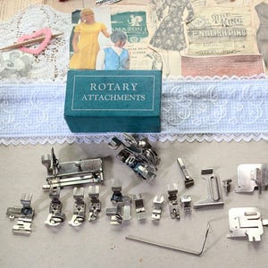 Puede incluir: Una caja rectangular de color verde azulado con la etiqueta ROTARY ATTACHMENTS en texto blanco, acompañada de una colección de varios prensatelas y accesorios de metal plateado para máquinas de coser, incluyendo una guía metálica fina.