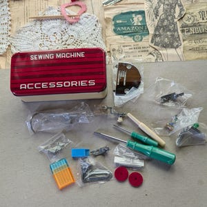 May include: A collection of sewing machine accessories, including a red and white metal box labeled "SEWING MACHINE ACCESSORIES." Various tools, bobbins, and presser feet are scattered around the box, along with vintage paper ephemera.