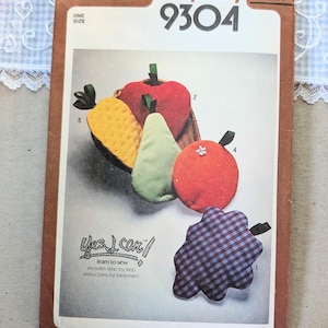 May include: A Simplicity 9304 sewing pattern envelope featuring illustrations of five fabric fruit and vegetable shapes including a red apple, a green pear, an orange, a yellow textured piece, and a blue and black checkered star-like shape.
