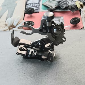 May include: A close-up of a silver sewing machine foot. The foot has multiple parts, including a lever, a presser foot, and a screw. The background includes pieces of paper and small black objects.