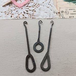May include: Three antique metal lace tools with hook ends and loop handles. The handles are stamped with text, including "Polish Laces" and "E.L. Troy Main St, Kill Haven, PA."