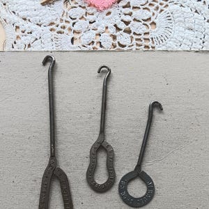 May include: Three vintage metal shoe button hooks of varying sizes. The hooks are dark gray with a curved hook at the top and a loop at the bottom. A pink heart-shaped decoration and a wooden knitting needle are in the background.