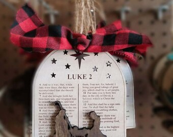 May include: A wooden Christmas ornament shaped like a bell with a red and black plaid ribbon. The ornament features a cutout of a shepherd and a star with the text "LUKE 2" and a portion of the Bible verse Luke 2:6-13.