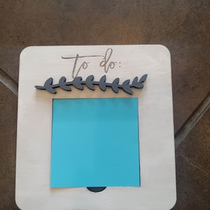 May include: White wooden to-do list holder with a black leaf design and a blue sticky note.