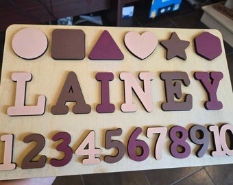 May include: A wooden puzzle with the name "LAINEY" spelled out in pink, brown, and purple letters. The puzzle also includes a variety of shapes, including a circle, square, triangle, heart, star, and octagon. The numbers 1 through 10 are also included in the puzzle.
